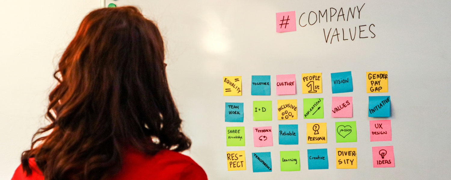 From posters to practice: How to bring your organization's values to life - Photo by Walls.io on Unsplash (A woman standing in front of a white board with company values written on it)